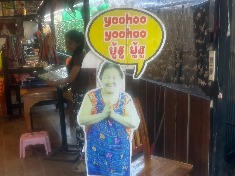 A life-sized cardboard cut-out of Madame Yoohoo with a yellow speech bubble reading "yoohoo yoohoo" inside her restaurant on Koh Lipe